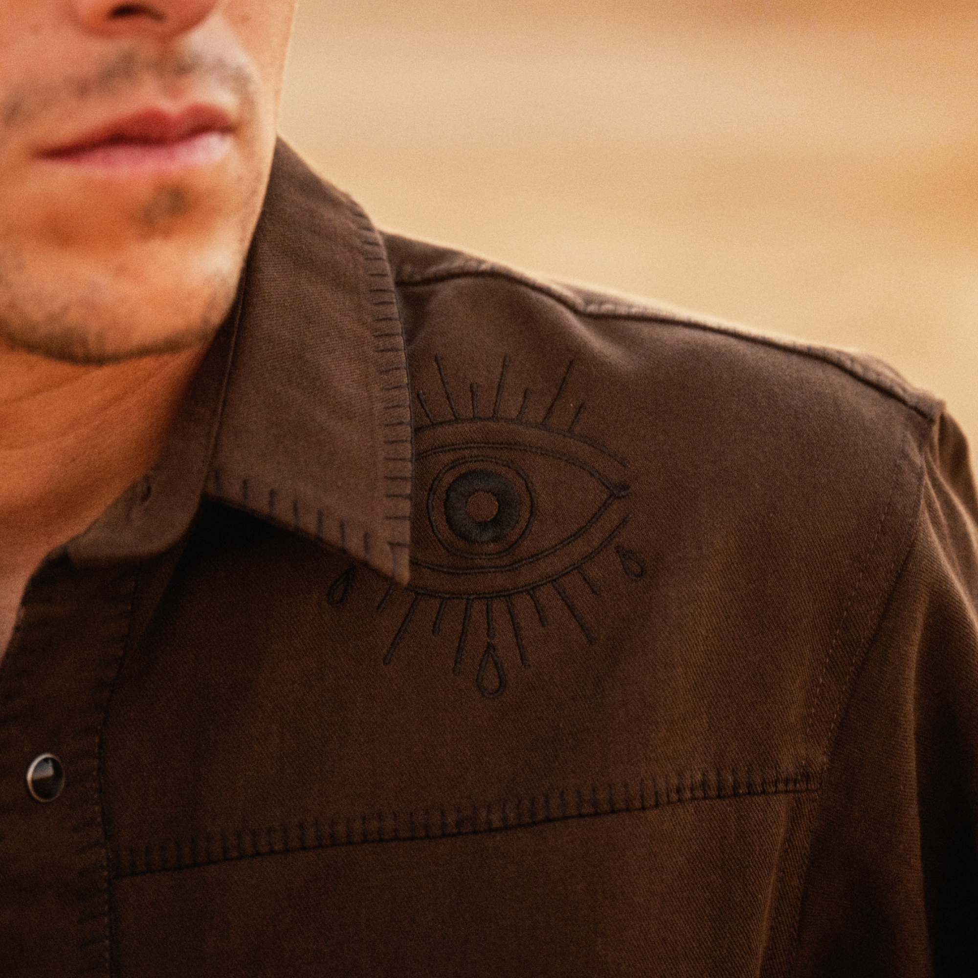 Brown button-up shirt with embroidered eye design on chest pocket, worn by man against neutral background